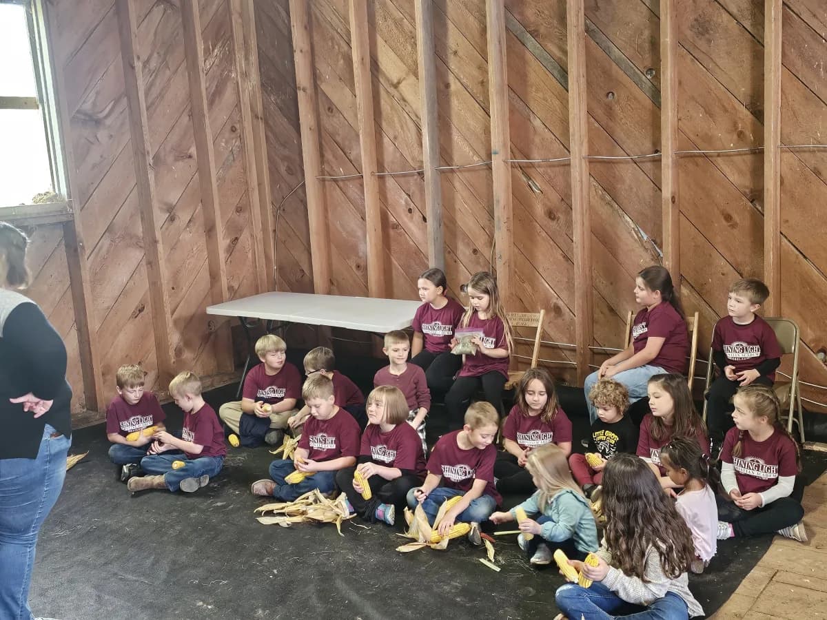 Students in school shirts learning about corn husking in a barn